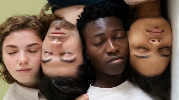 Two people wearing casual clothing are closely positioned against a light green background, with one person’s hand gently resting on the other’s shoulder.