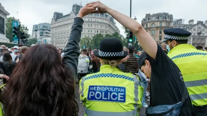 Two Metropolitan Police officers in high-visibility jackets stand among a crowd while a person raises clasped hands above their head during a street demonstration in an urban setting.