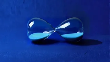 A glass hourglass lies horizontally on a deep blue surface, with blue sand evenly distributed between its two bulbs.