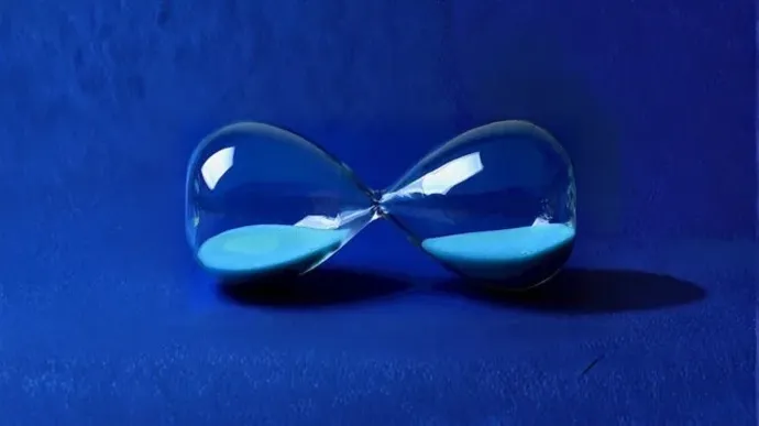 A glass hourglass lies horizontally on a deep blue surface, with blue sand evenly distributed between its two bulbs.