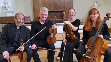 Four musicians seated, holding string instruments, smiling at the camera. They are in a room with wooden flooring and a wooden backdrop.