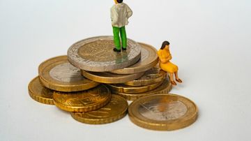 Miniature figures on stacked coins; a man stands on top, and a woman sits on the edge, depicting financial inequality.