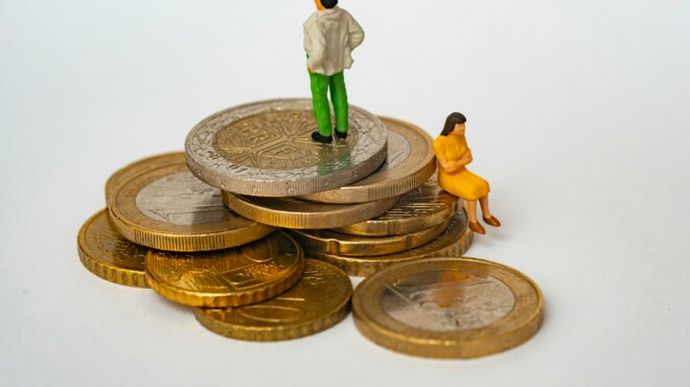 Miniature figures on stacked coins; a man stands on top, and a woman sits on the edge, depicting financial inequality.
