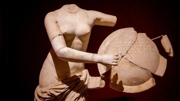 Headless marble statue holding a broken, inscribed shield against a dark background.