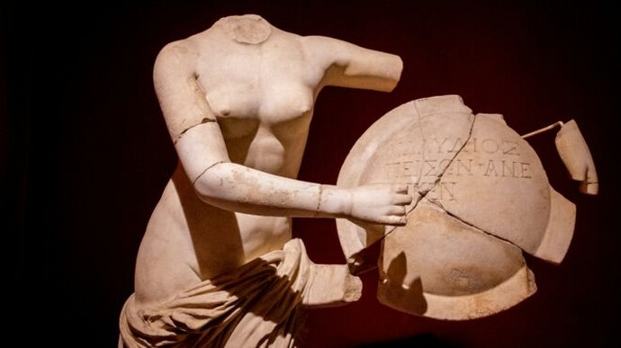 Headless marble statue holding a broken, inscribed shield against a dark background.