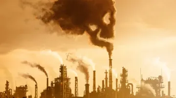A row of industrial chimneys releases thick black smoke into the sky, creating a dramatic scene of heavy air pollution against an orange-tinted background.