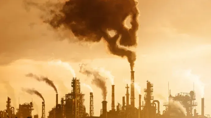 A row of industrial chimneys releases thick black smoke into the sky, creating a dramatic scene of heavy air pollution against an orange-tinted background.