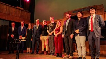 A group of ten people dressed in formal attire stand on a stage, smiling, with a wooden piano and a green wall in the background.