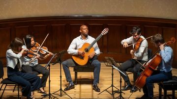 A quintet performs on stage with a guitarist, two violinists, a violist, and a cellist, all seated and focused on their music.