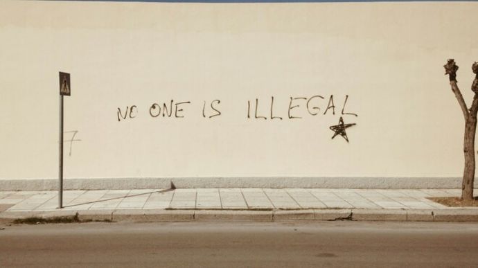 Graffiti on a beige wall reads "NO ONE IS ILLEGAL" with a star symbol, beside a leafless tree and a pedestrian crossing sign.