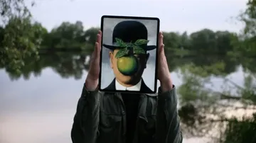 A person holds a tablet in front of their face displaying René Magritte’s painting of a man in a bowler hat with a green apple covering his face, blending art with the outdoor background.