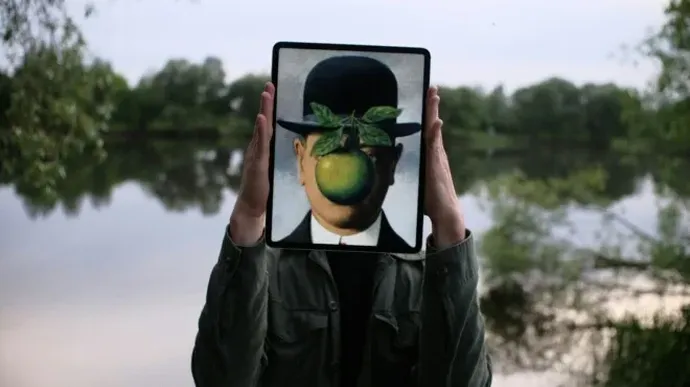 A person holds a tablet in front of their face displaying René Magritte’s painting of a man in a bowler hat with a green apple covering his face, blending art with the outdoor background.
