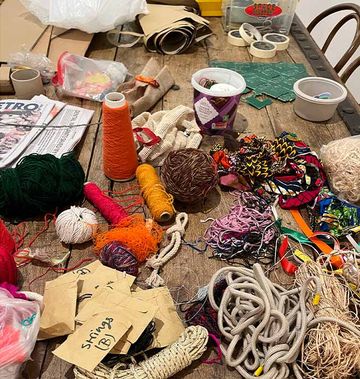 A cluttered table with colorful yarn, strings, fabric scraps, tape, and crafting materials scattered around.