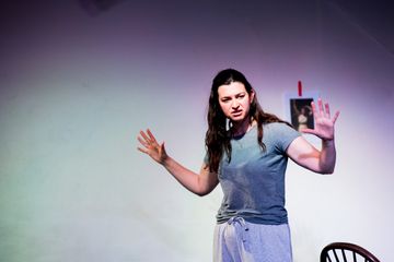 Person performing on stage, raising hands, wearing casual clothes, spotlight lighting, simple backdrop and chair