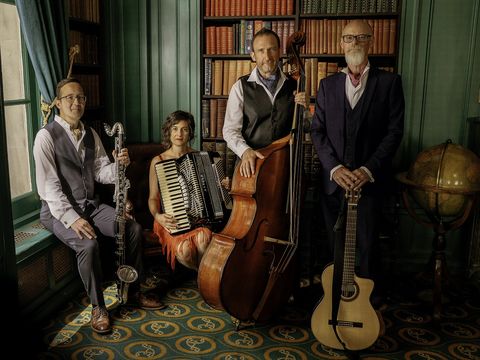 Four musicians in vintage attire pose in a library, holding instruments including a clarinet, accordion, double bass, and guitar.