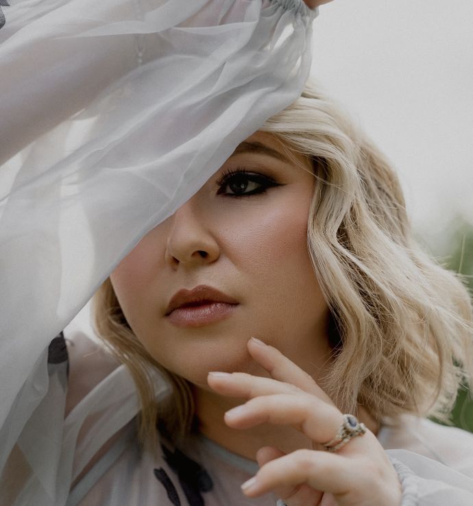 Close up of blonde woman with hand against chin holding chiffon sleeve over one side of face