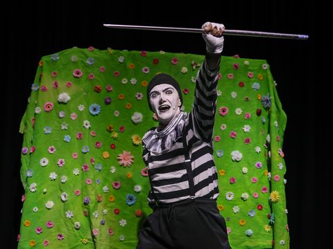 Mime artist in black and white costume performing against a vibrant green floral backdrop, holding a staff with an expressive pose.