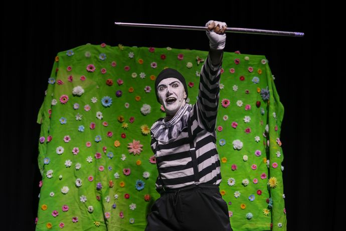 Mime artist in black and white costume performing against a vibrant green floral backdrop, holding a staff with an expressive pose.