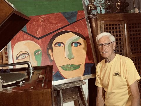 Person standing beside colourful abstract portrait artwork, vintage gramophone, wooden cabinet, and artist's easel indoors.