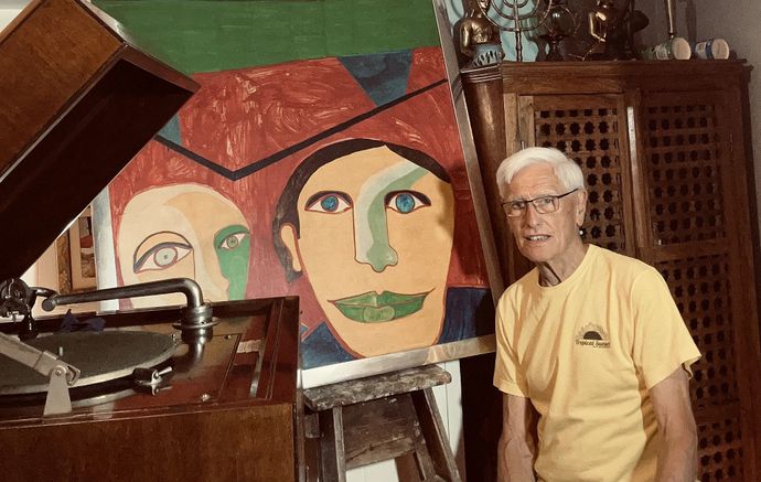 Person standing beside colourful abstract portrait artwork, vintage gramophone, wooden cabinet, and artist's easel indoors.