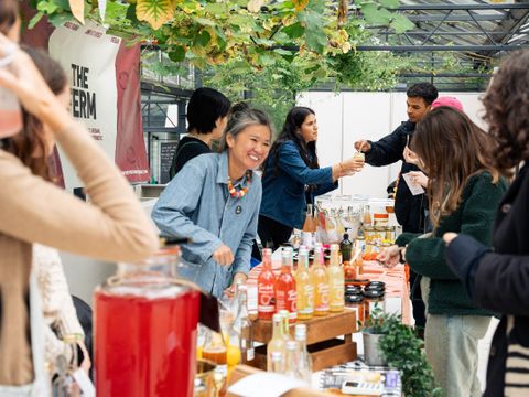 People at a market stall with bottles of drinks and jars on display under overhead plants in a greenhouse setting.