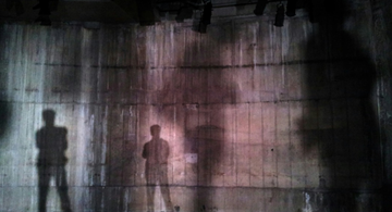 Shadows of two people projected on a textured, dimly lit wall beneath stage lights, creating a mysterious and artistic scene.