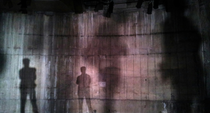Shadows of two people projected on a textured, dimly lit wall beneath stage lights, creating a mysterious and artistic scene.