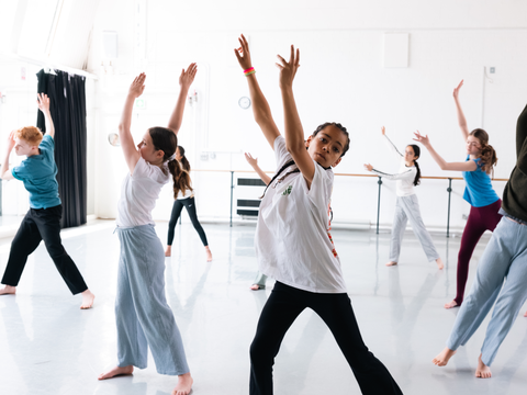 Children in a dance studio practicing a routine, with arms raised and expressing movement, wearing casual attire.