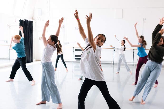 Children in a dance studio practicing a routine, with arms raised and expressing movement, wearing casual attire.