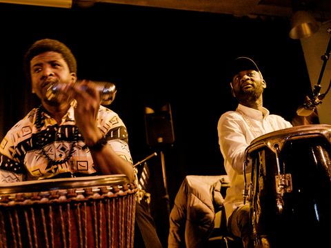 Two percussionists performing on stage with drums, microphones, warm lighting, and live music.