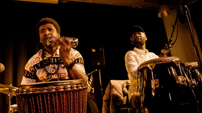 Two percussionists performing on stage with drums, microphones, warm lighting, and live music.