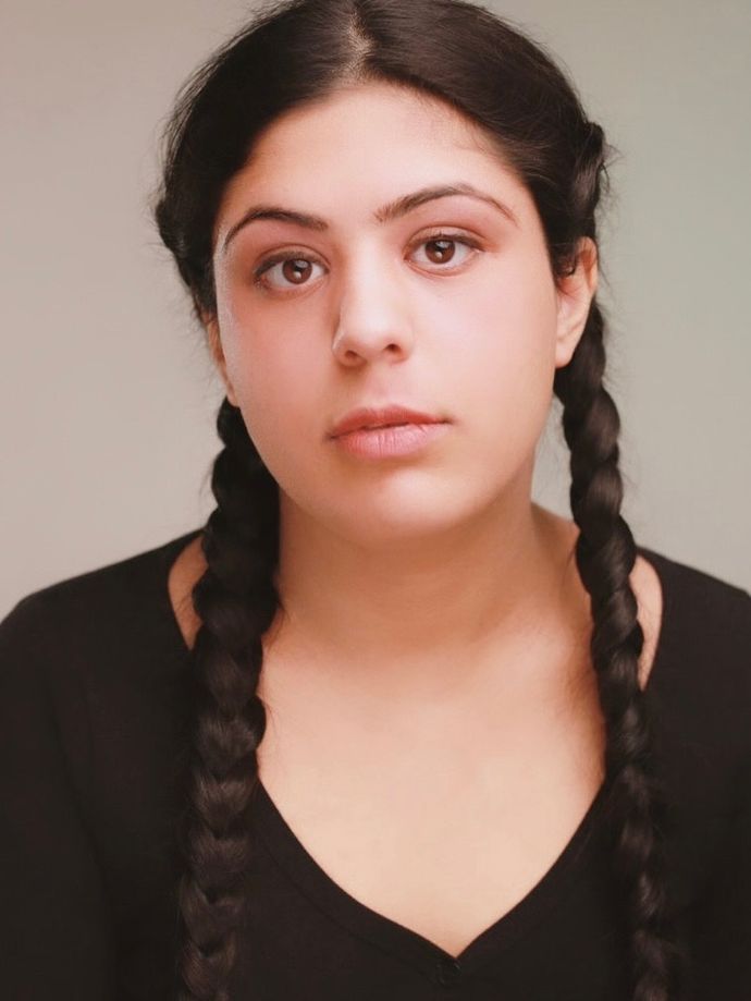 Scarlett Westbrook with long dark braids, wearing a black top, looks directly at the camera against a plain background.