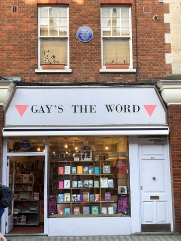Front view of "Gay's The Word" bookstore with a white facade displaying books in the window. Brick walls and upstairs windows in the background.