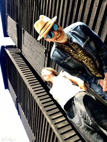 Two people standing outdoors in front of a tall, dark wooden structure. One person wears a straw hat, sunglasses, and a denim jacket, while the other wears a light top and dark pants. The photo is taken from a low angle, emphasizing the height of the building