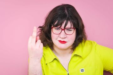 A person wearing red glasses raises a middle finger, displaying defiance against a bright pink background confidently.