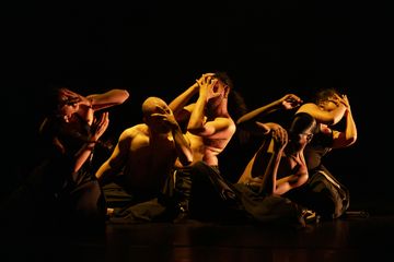 Five dancers in dynamic poses on a dimly lit stage, each with hands near face, creating a dramatic and expressive scene.
