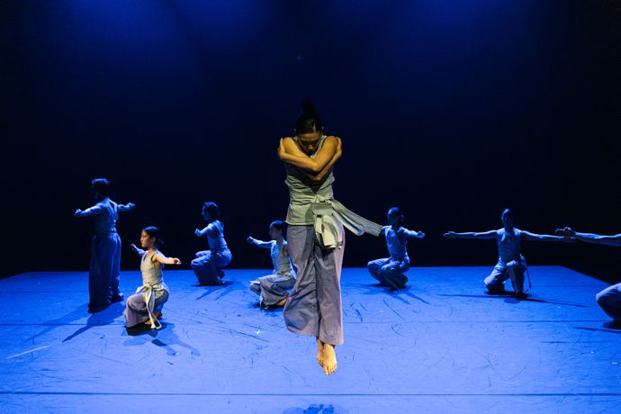 Dancer mid-air on stage, surrounded by others kneeling and reaching outward under blue lighting.
