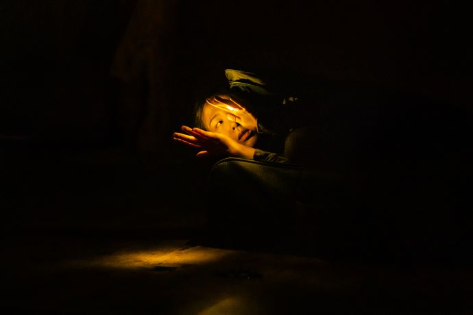 A person lies on a couch in dim light, their face illuminated dramatically, with a hand raised to their face, creating a mysterious atmosphere.