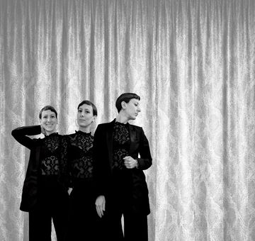 Three women in matching black outfits with lace tops stand in front of a textured curtain, each striking a different pose.