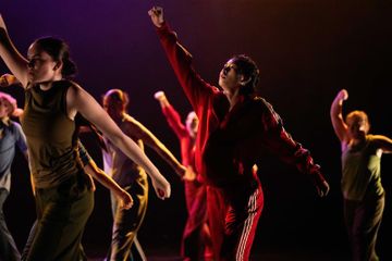 A group of dancers in dynamic poses, wearing varied athletic wear, perform under warm stage lighting.