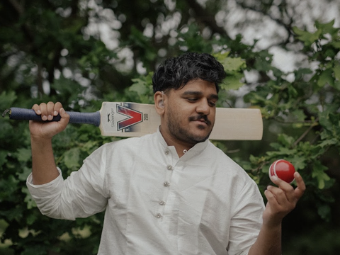 Person in a white shirt holding a cricket bat over their shoulder and looking at a red cricket ball held in their hand, standing outdoors by green foliage.