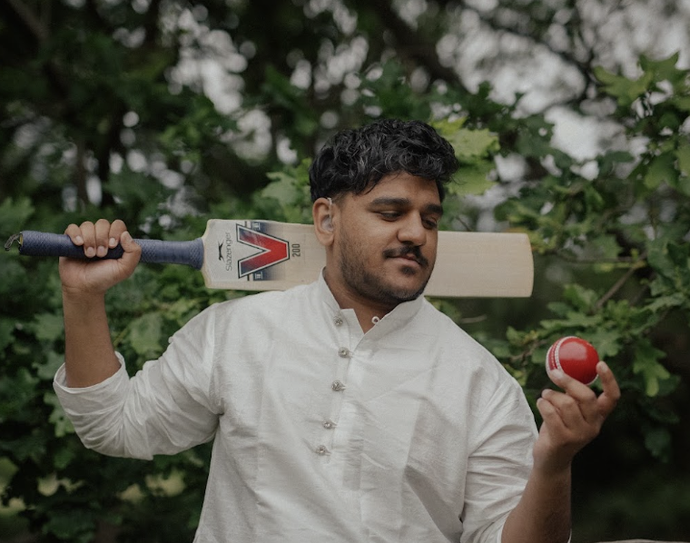 Person in a white shirt holding a cricket bat over their shoulder and looking at a red cricket ball held in their hand, standing outdoors by green foliage.