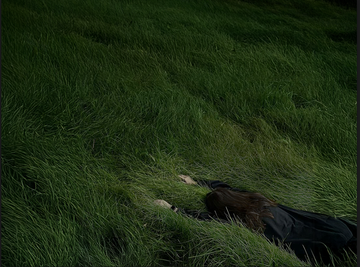 Person lying face down in tall, lush green grass, wearing a dark coat, blending into the natural surroundings.