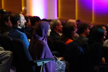 Audience seated in a dimly lit auditorium, watching a presentation, with colorful lights illuminating the background.