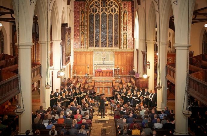 Choir and small orchestra performing at the front of a large, ornate church, with an audience seated in pews facing a grand altar and tall stained‑glass window