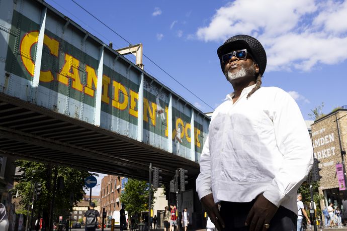 Jazzie B stands confidently under a bridge with "Camden" painted on it, against a blue sky with scattered clouds.
