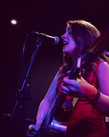 A person wearing a sleeveless red outfit is playing an electric guitar and singing into a microphone on a dimly lit stage with purple lighting.