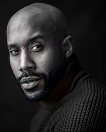 Black and white portrait of a bald man with a beard, wearing a textured sweater, looking intently at the camera against a dark background.