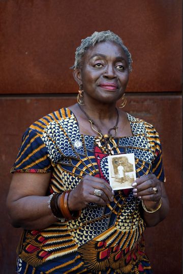 Elderly woman in vibrant patterned dress holds a vintage photo, standing against a rust-colored background.