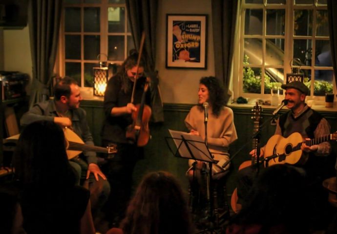 Four musicians — violinist, two guitarists and a singer — perform in a cozy, dimly lit room to a small audience.